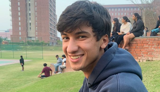 Smiling person in a blue hoodie sitting on grass, with people and buildings in the background.