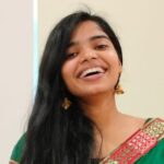 Smiling woman in a green traditional outfit with gold earrings, standing indoors.