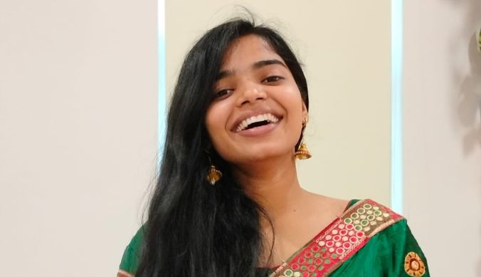 Smiling woman in a green traditional outfit with gold earrings, standing indoors.