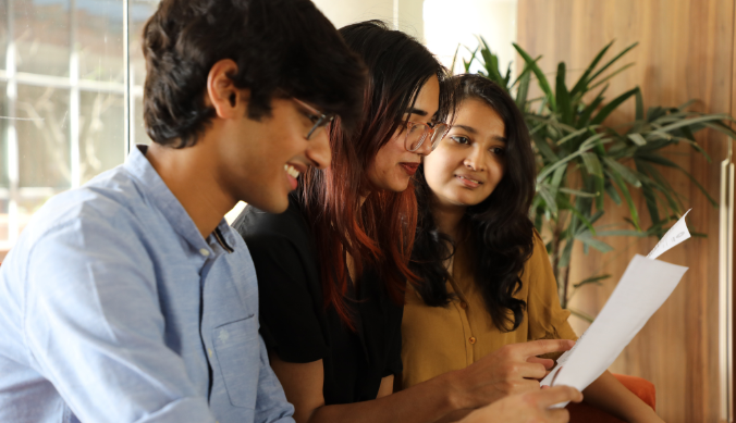 Three people sitting together, reviewing a document, with a plant and wooden wall in the background.