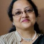 Woman wearing glasses, a striped blouse, and a black saree with a pearl necklace, smiling gently.