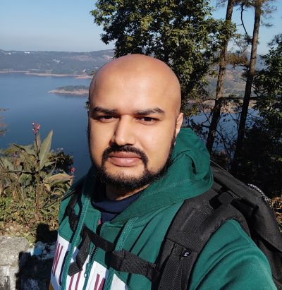 Man in green hoodie and backpack standing outdoors with trees and a lake in the background.