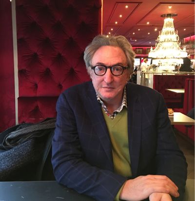 Man with glasses seated at a table in a red, elegant restaurant with a chandelier in the background.