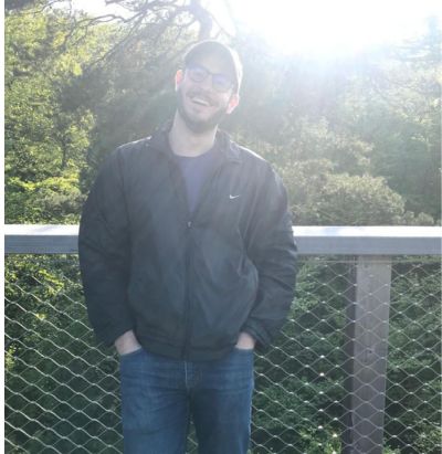 Man in a black jacket and cap smiling on a bridge with sunlight and trees in the background.
