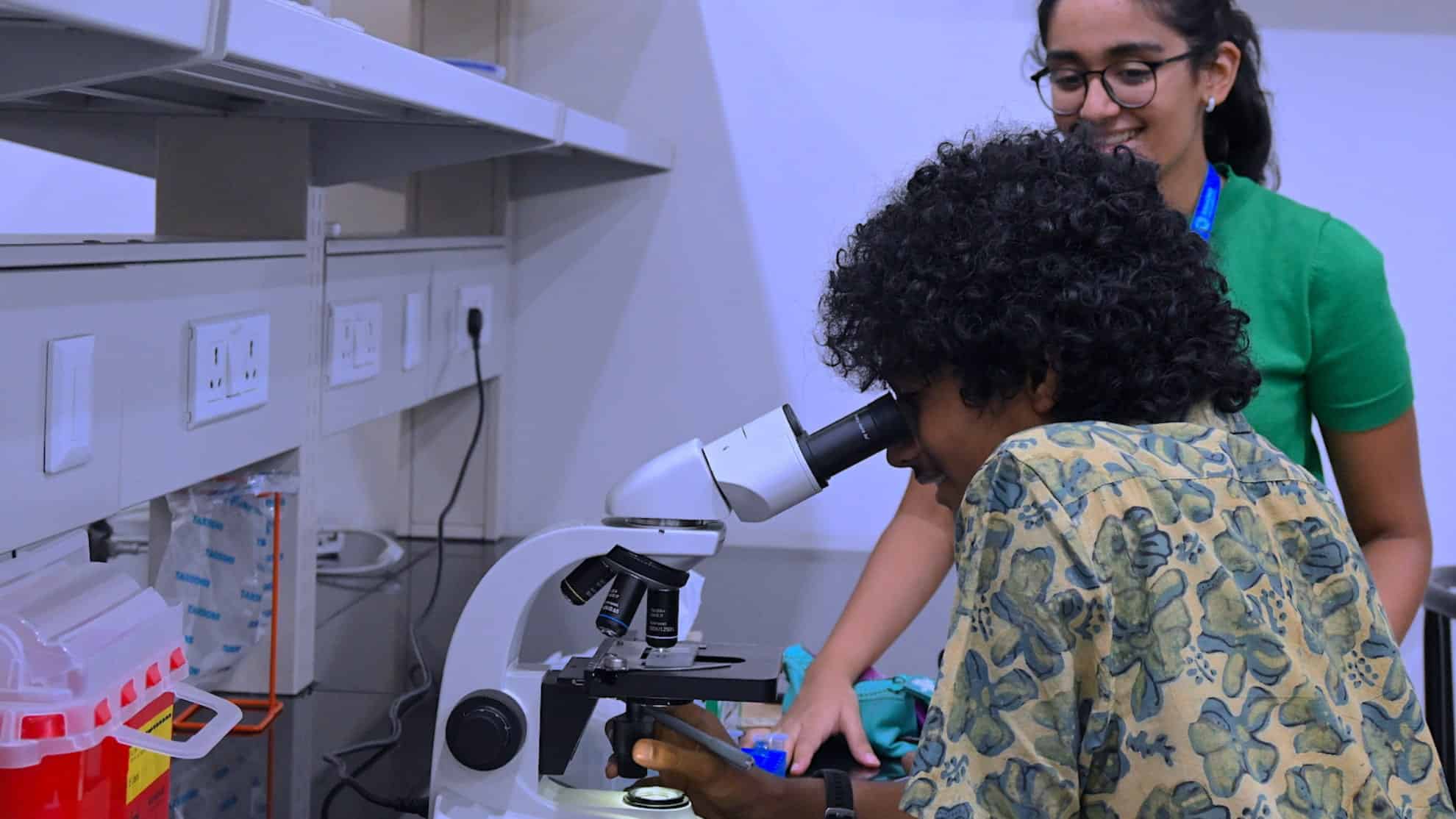 Person using a microscope in a lab while another person smiles and observes.