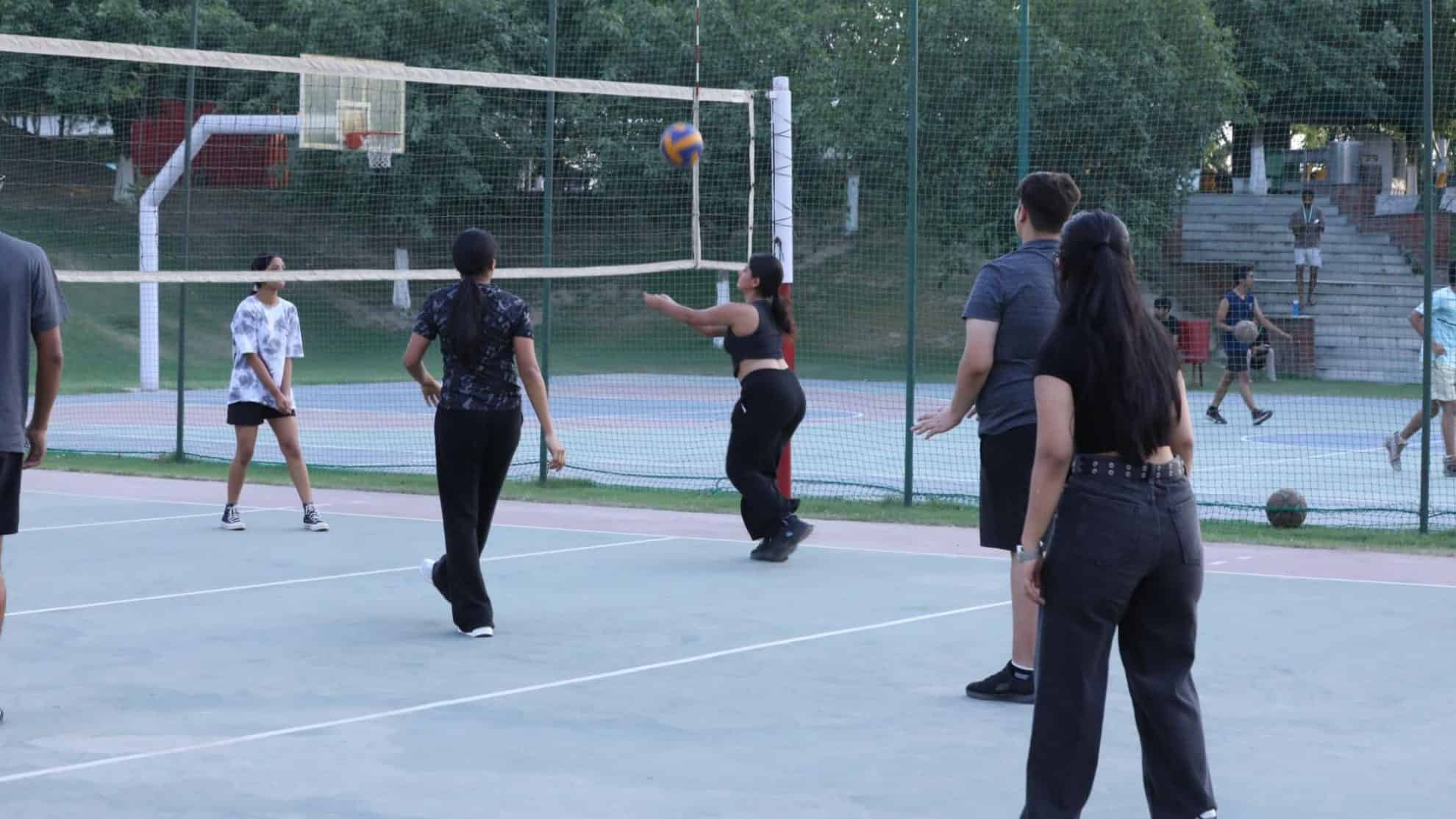 People playing volleyball on an outdoor court, surrounded by greenery and a basketball court.