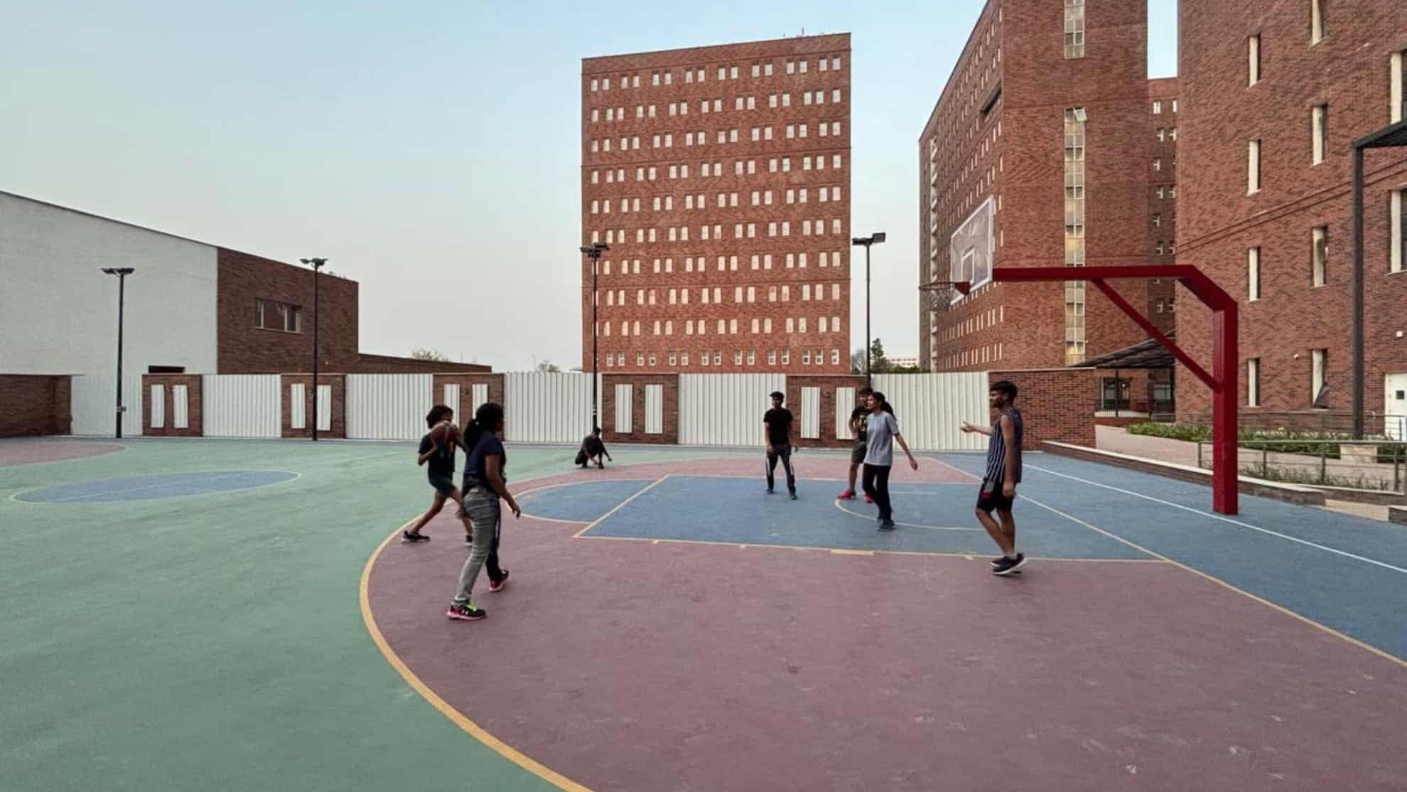 People playing basketball on an outdoor court surrounded by brick buildings and a clear sky.