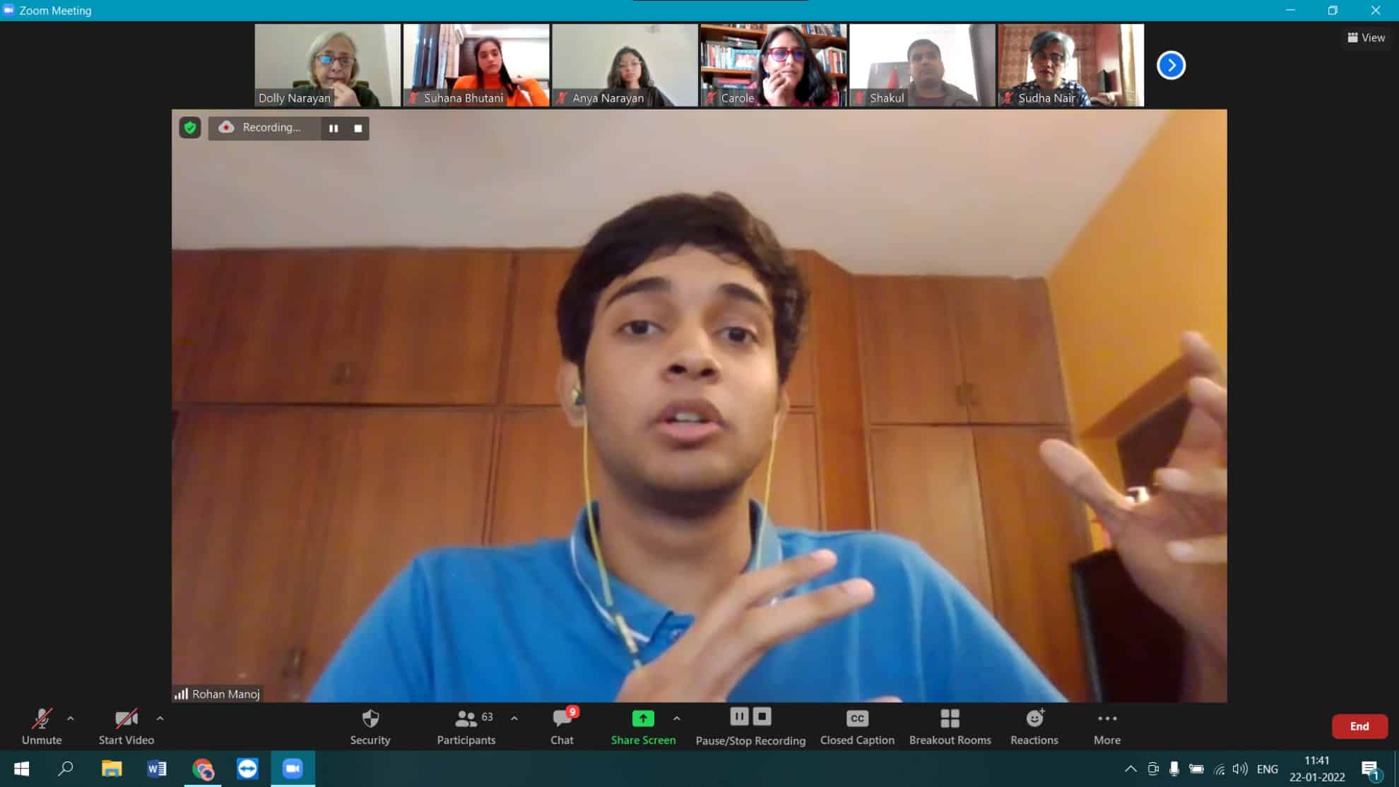 Person in a blue shirt speaking during a Zoom call with six participants visible at the top.