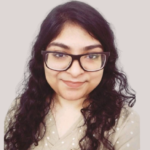 Woman with long curly hair, glasses, and a beige polka-dotted top, smiling against a plain background.