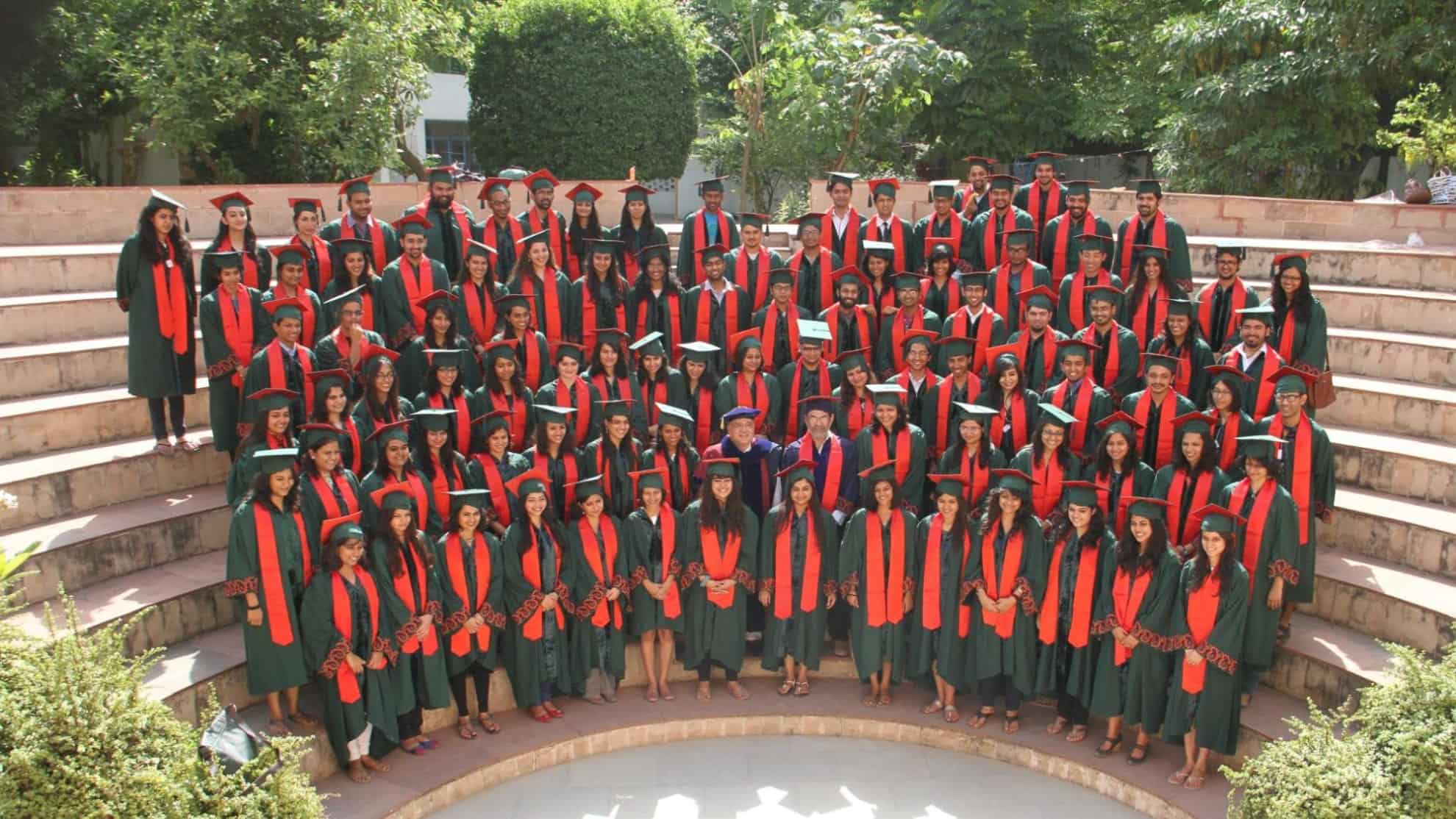 Graduates in green gowns and red stoles pose on amphitheater steps with trees in the background.