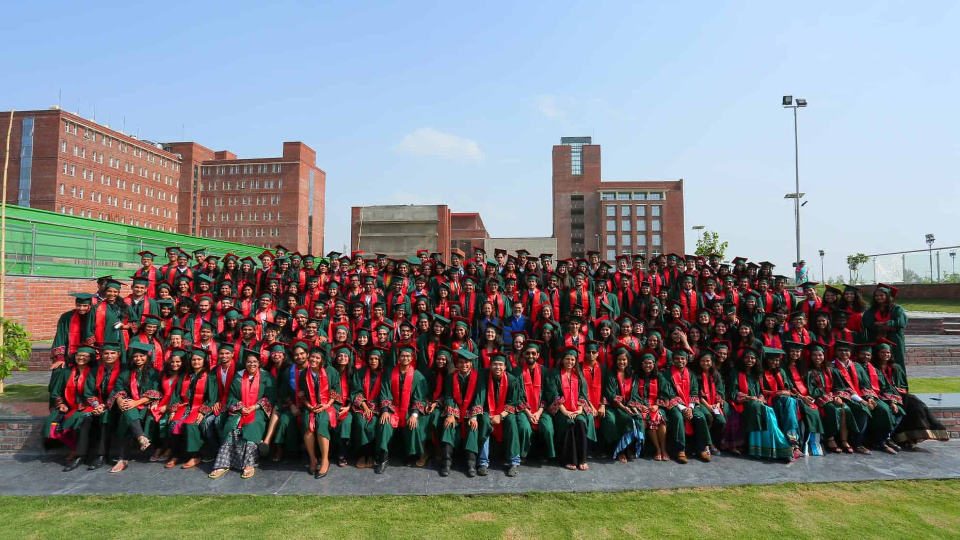 Graduates in green gowns and red stoles seated outdoors with brick buildings in the background.