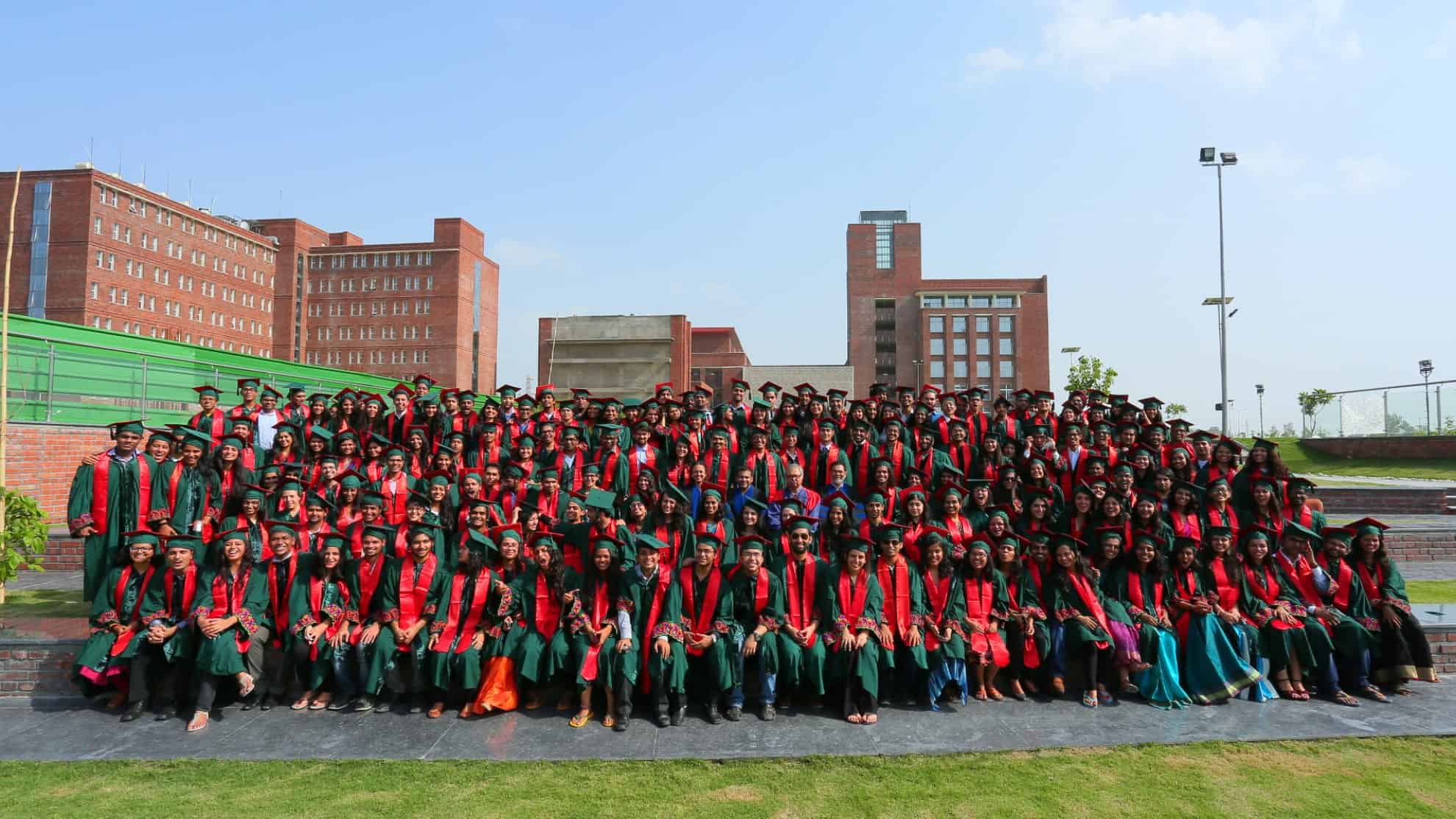 Graduates in green gowns and red stoles pose outdoors with a brick campus building in the background.