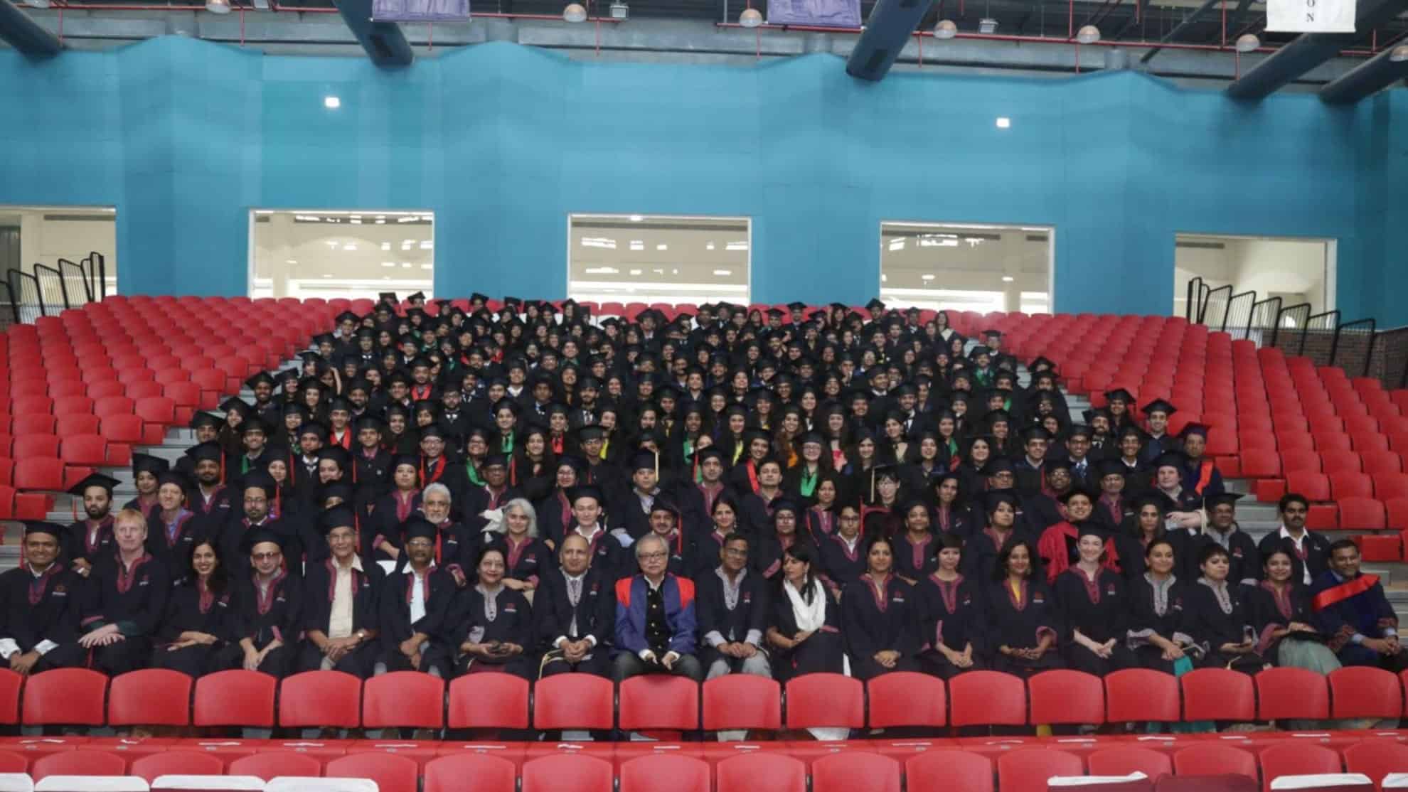 Graduates in caps and gowns seated in rows on red chairs, posing indoors with faculty members.