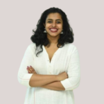 Smiling woman with curly hair wearing a white top, arms crossed, standing against a light background.