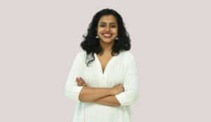Smiling woman with curly hair wearing a white top, arms crossed, standing against a light background.