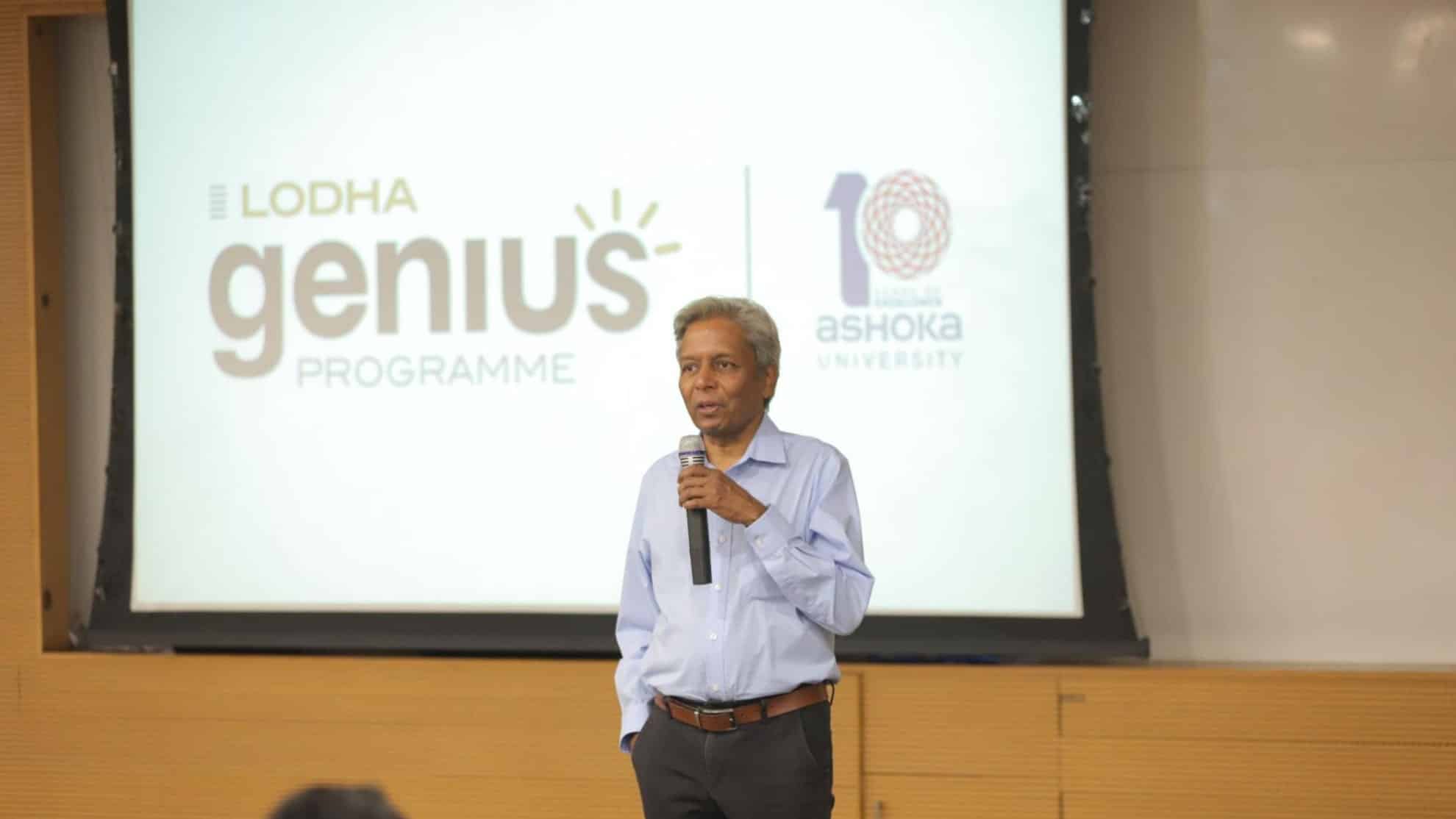 Man speaking on stage with a microphone, Lodha Genius Programme and Ashoka University logos behind him.