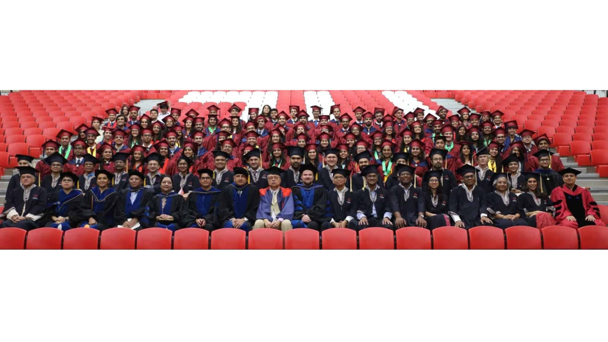 Graduates in maroon gowns and faculty in academic regalia seated in rows on red auditorium seats.