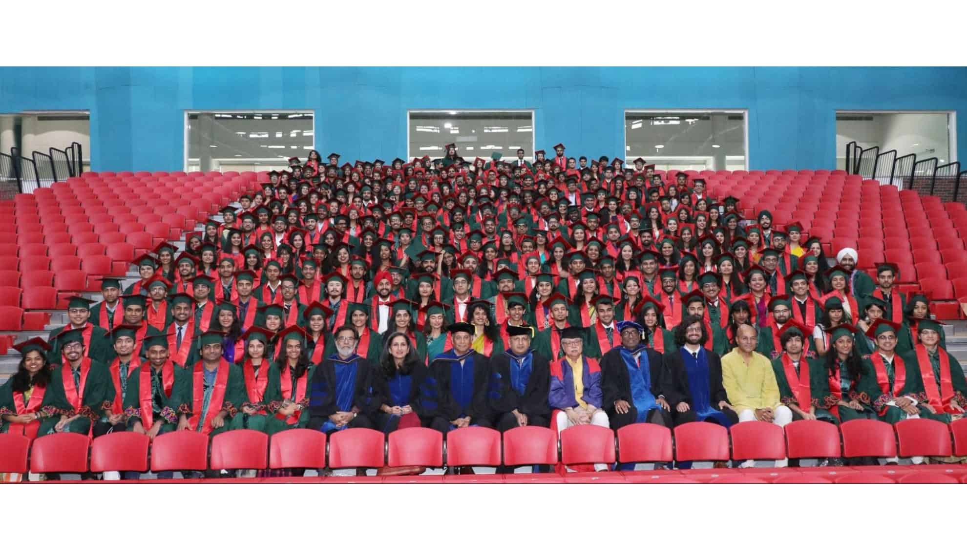 Graduates in green gowns with red stoles seated with faculty in a large auditorium.