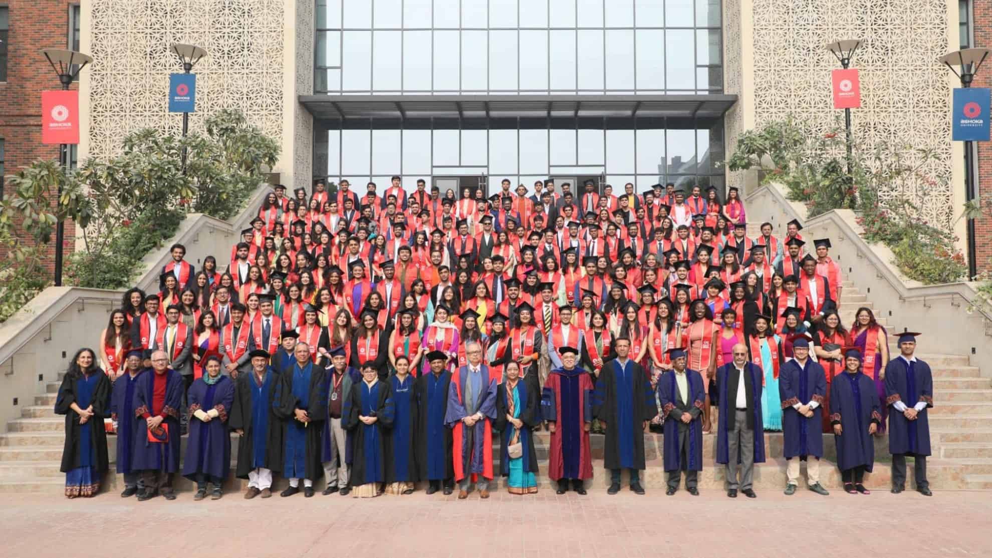 Graduates in red stoles and faculty in robes pose on stairs in front of a modern building.