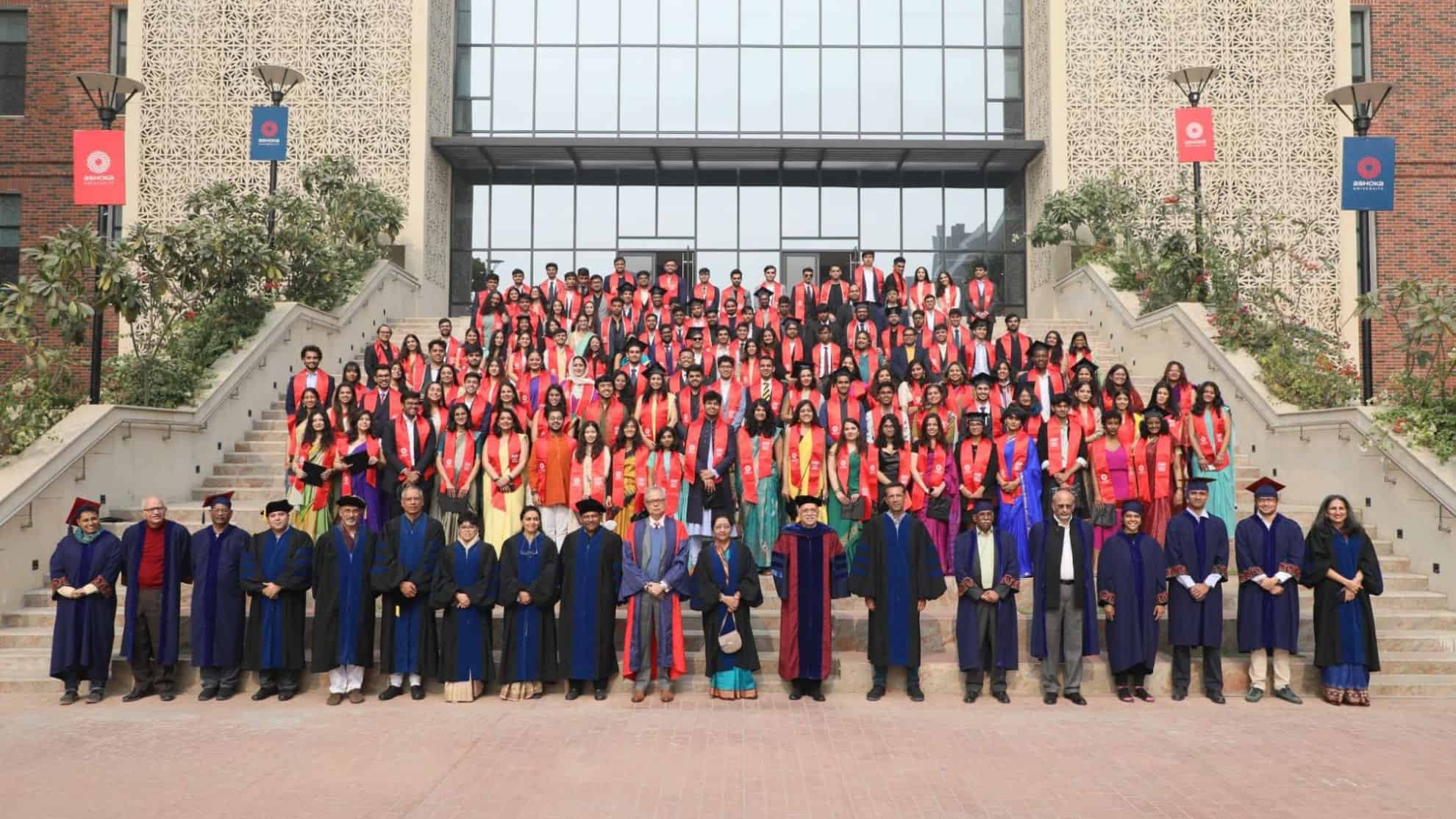 Graduates in red stoles and faculty in robes pose on steps in front of a modern building.