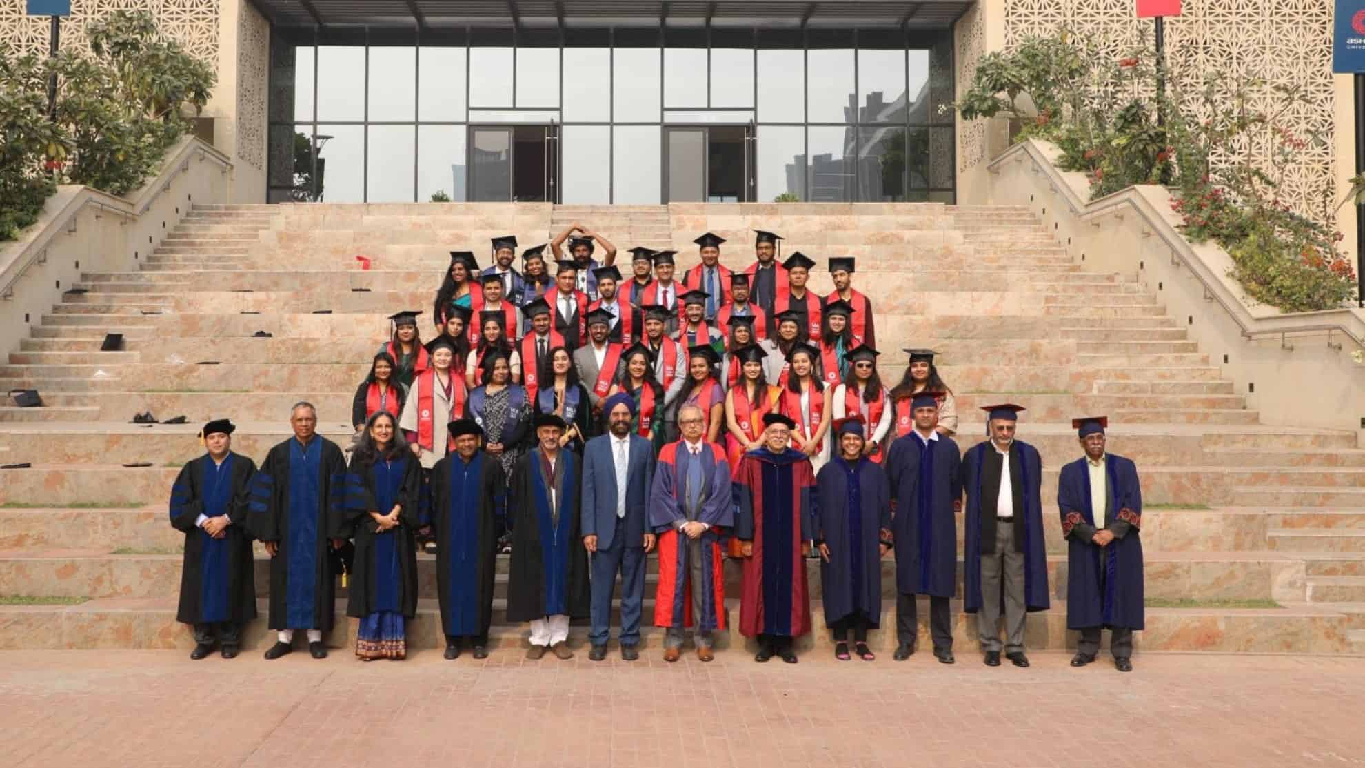 Graduates in caps and gowns with faculty in robes pose on steps in front of a modern building.