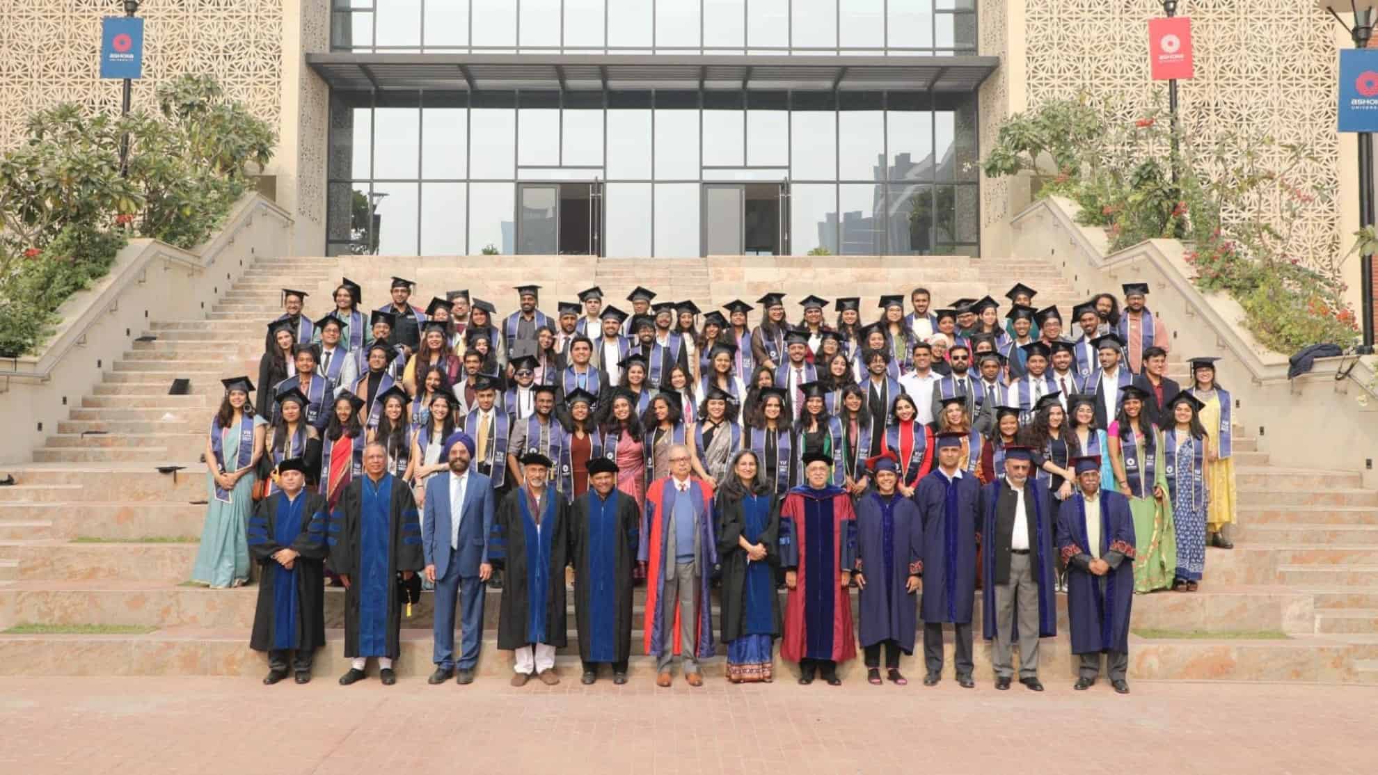 Graduates in caps and gowns with faculty on building steps, posing for a group photo outdoors.