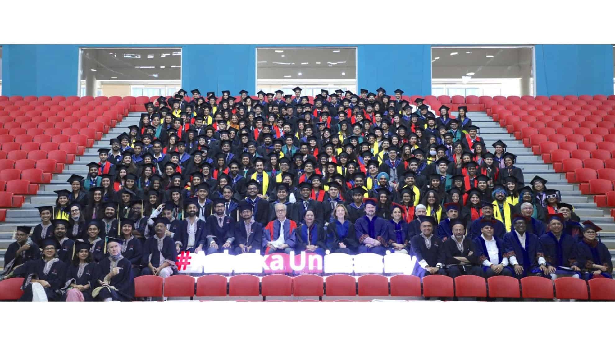Graduates in caps and gowns seated in rows with faculty members in front, red chairs in the background.