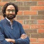 Person with curly hair, glasses, and a blue jacket standing confidently against a brick wall.