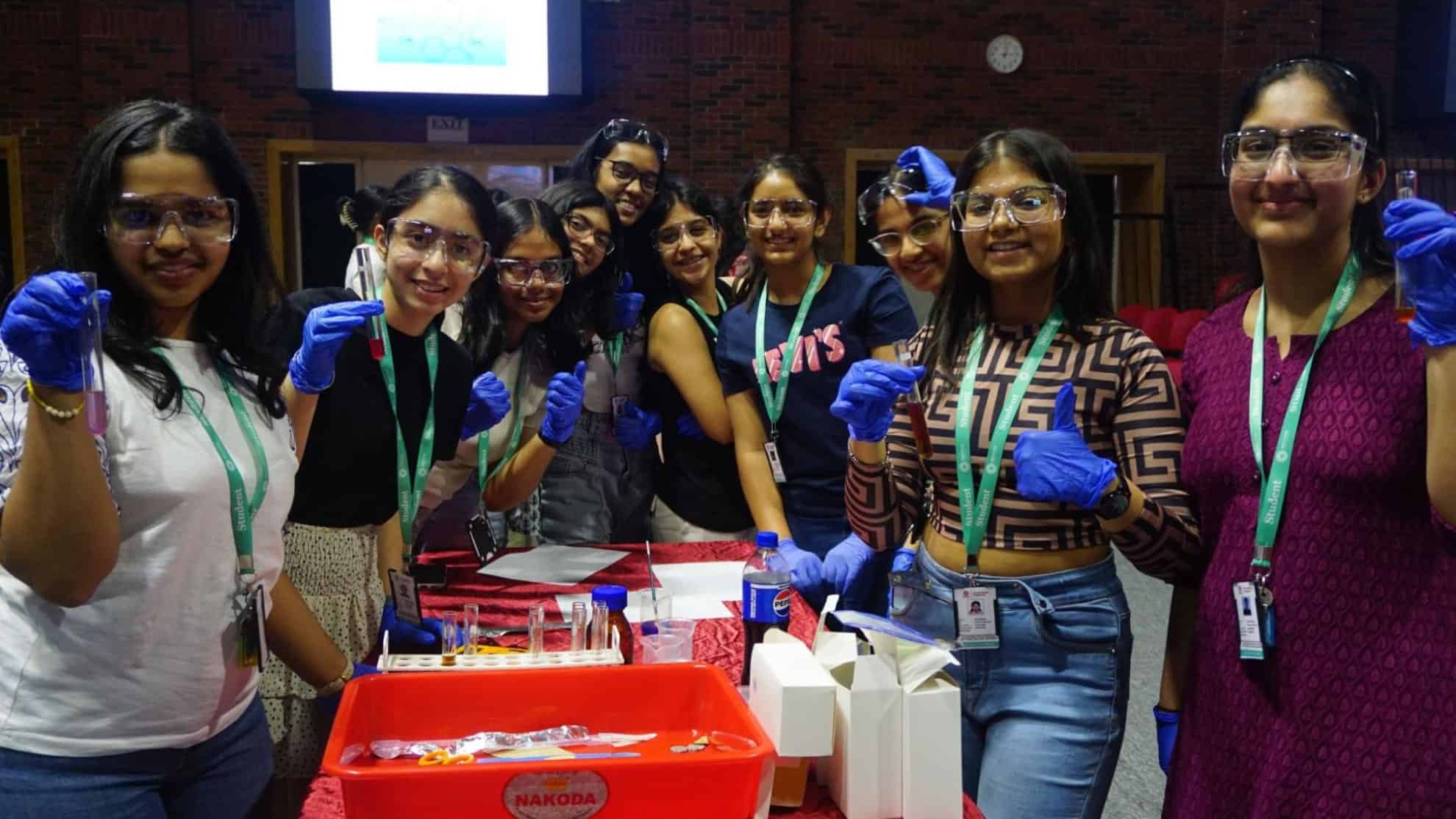 Group of students wearing gloves and goggles holding test tubes, smiling in a science activity setup.