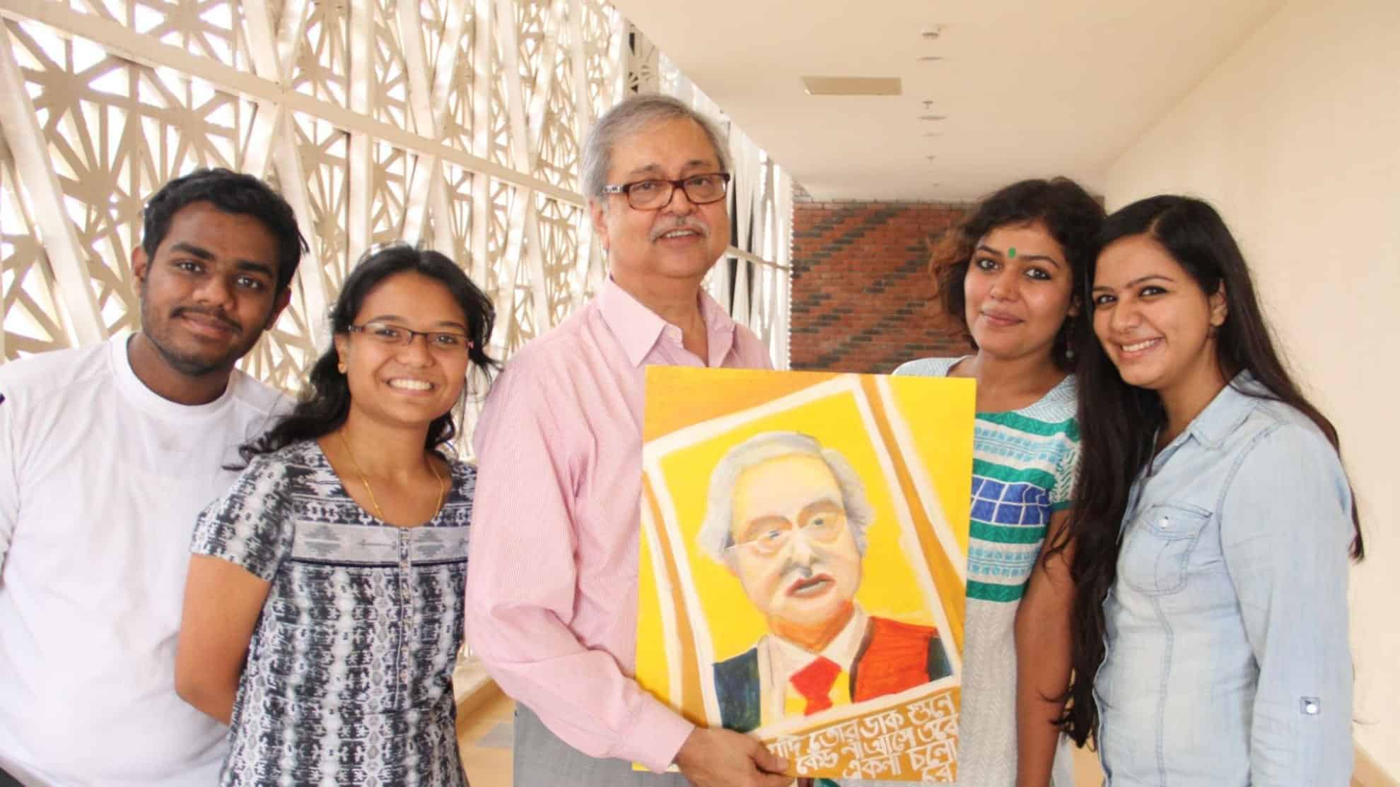Group of five smiling people holding a colorful portrait in a well-lit hallway.