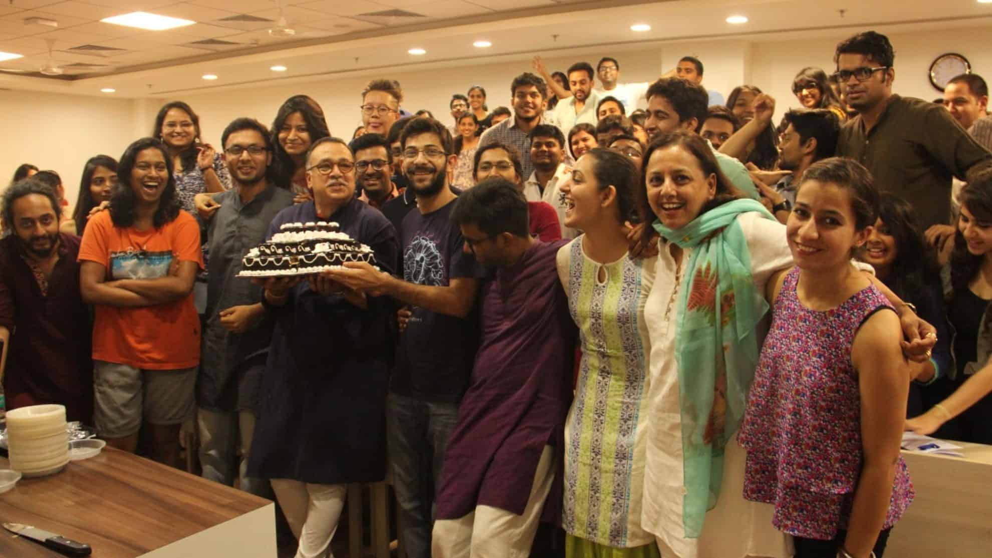 Group of people smiling, celebrating with a cake in a brightly lit room.