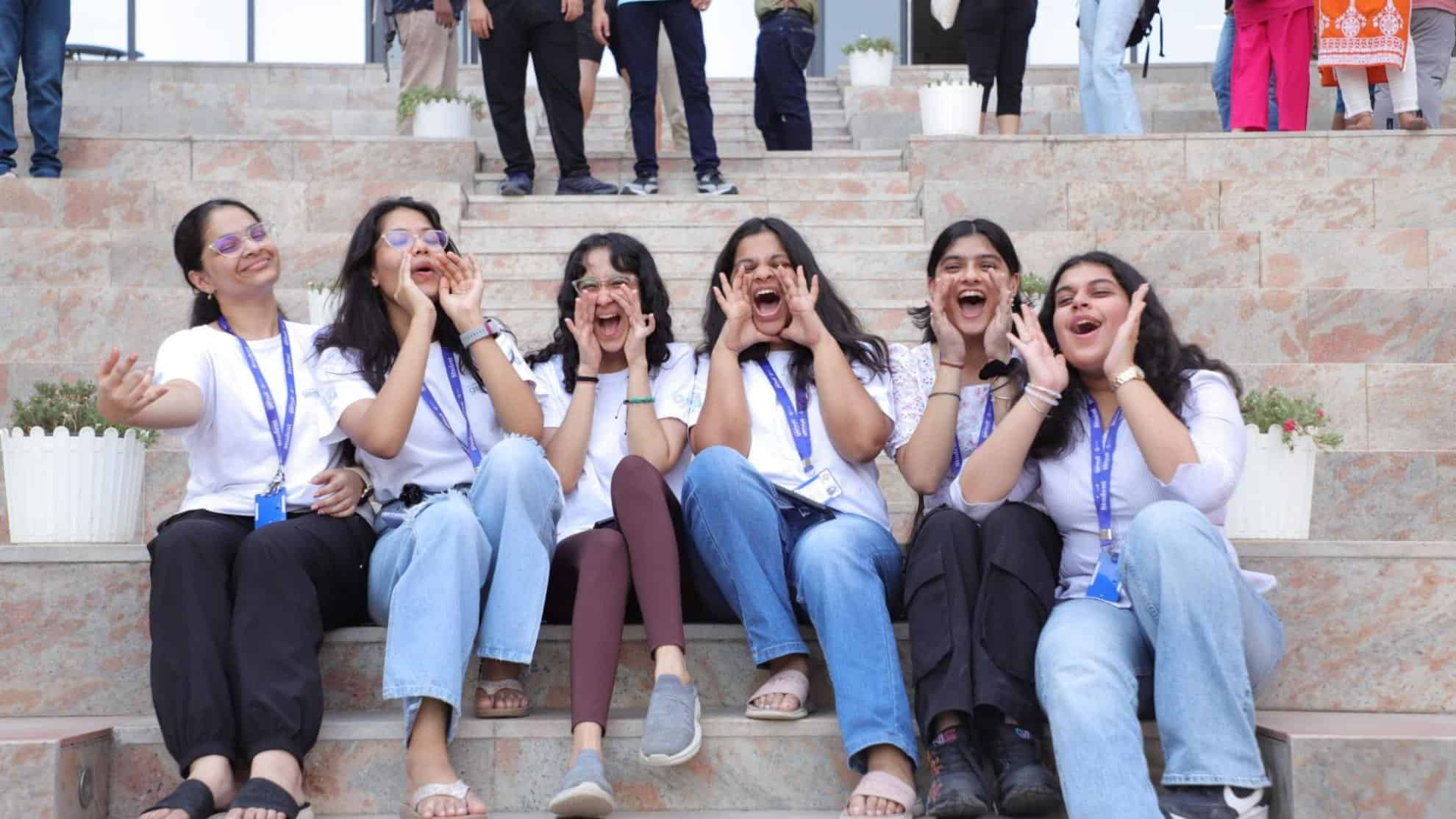 Six women sitting on steps, wearing white shirts with lanyards, laughing and cheering together.