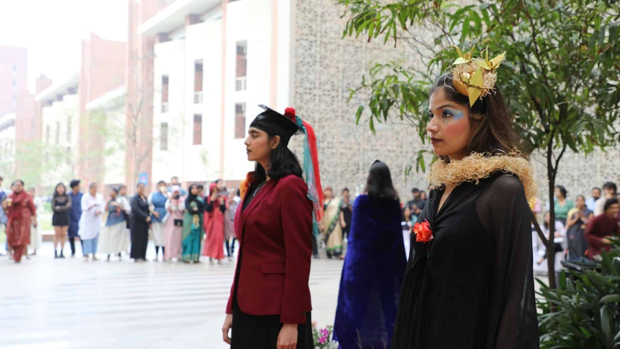 Two women in costumes stand outdoors, with a crowd and buildings in the background.