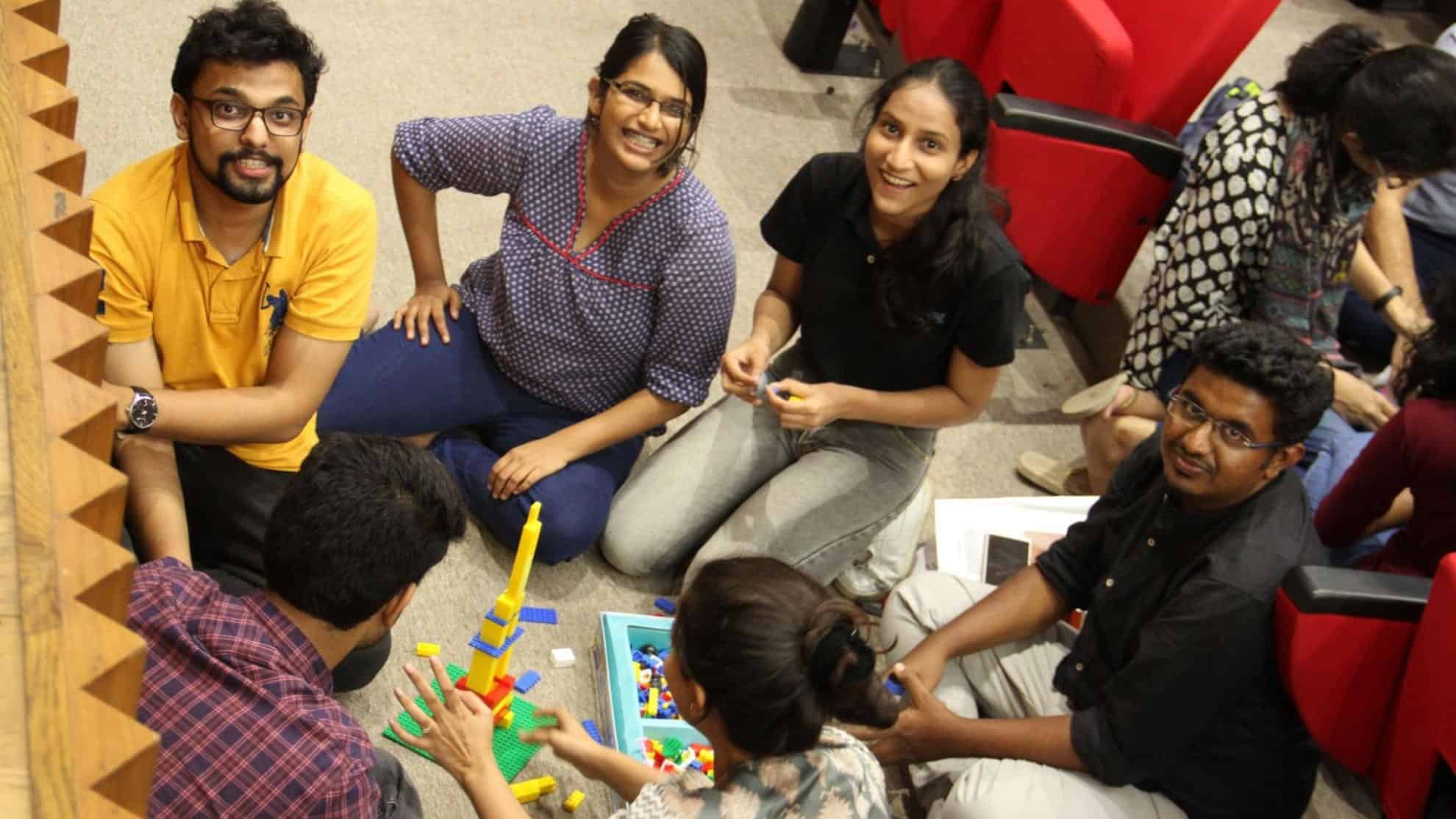 Group of six people seated on the floor, smiling and building with colorful Lego bricks.