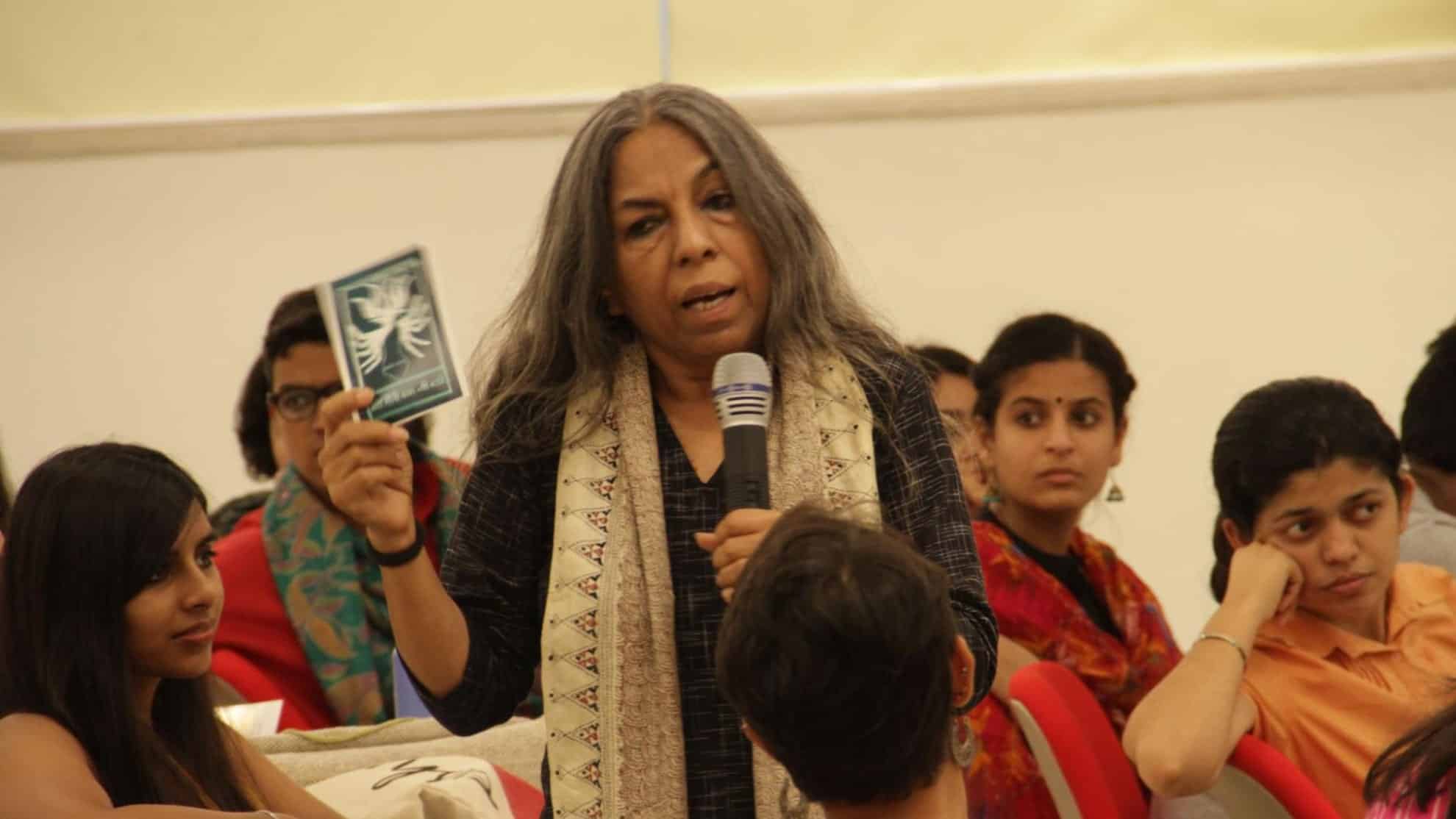 Woman holding a booklet and microphone, speaking to an audience seated in a classroom setting.