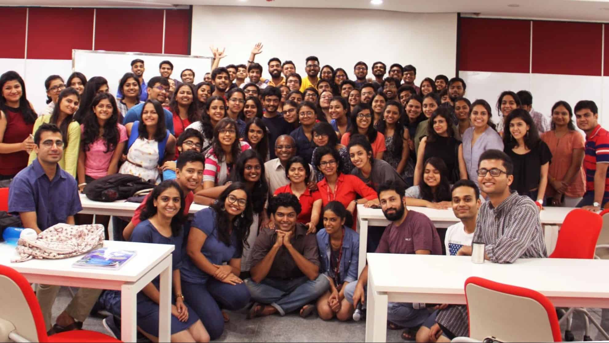 Group of smiling people in a classroom setting with whiteboards and red chairs in the background.