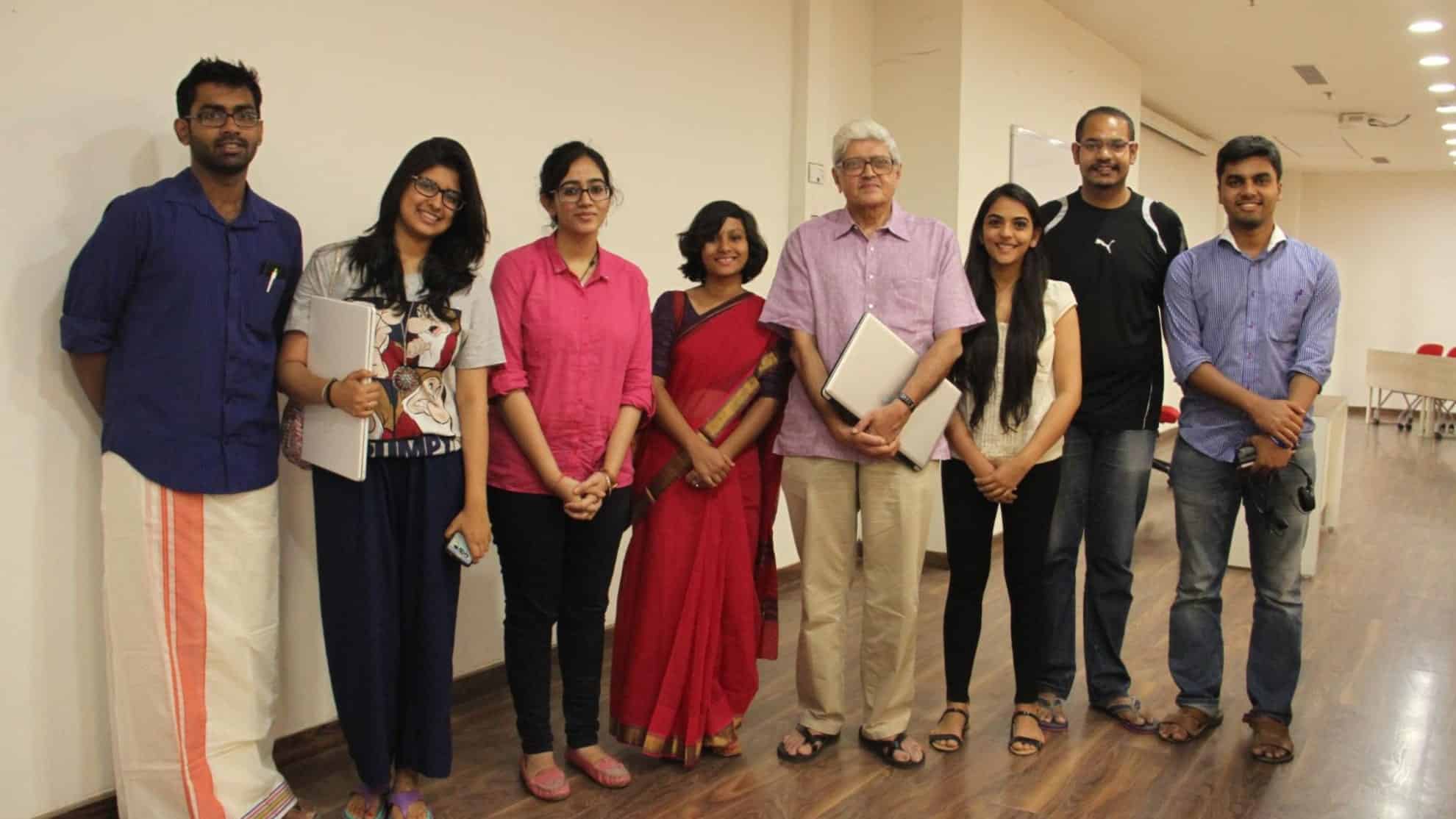 Group of eight people standing indoors, some holding laptops, smiling, casual and formal attire.