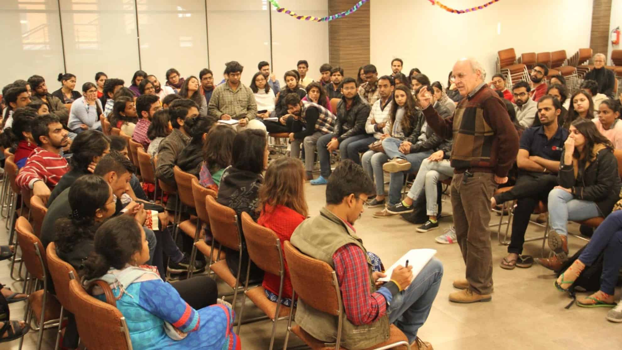 A man speaks to a seated group in a classroom setting, with attendees listening attentively.