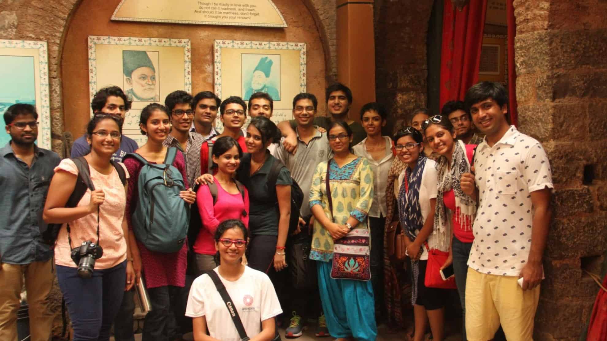 Group of young adults smiling indoors with framed artwork and stone walls in the background.