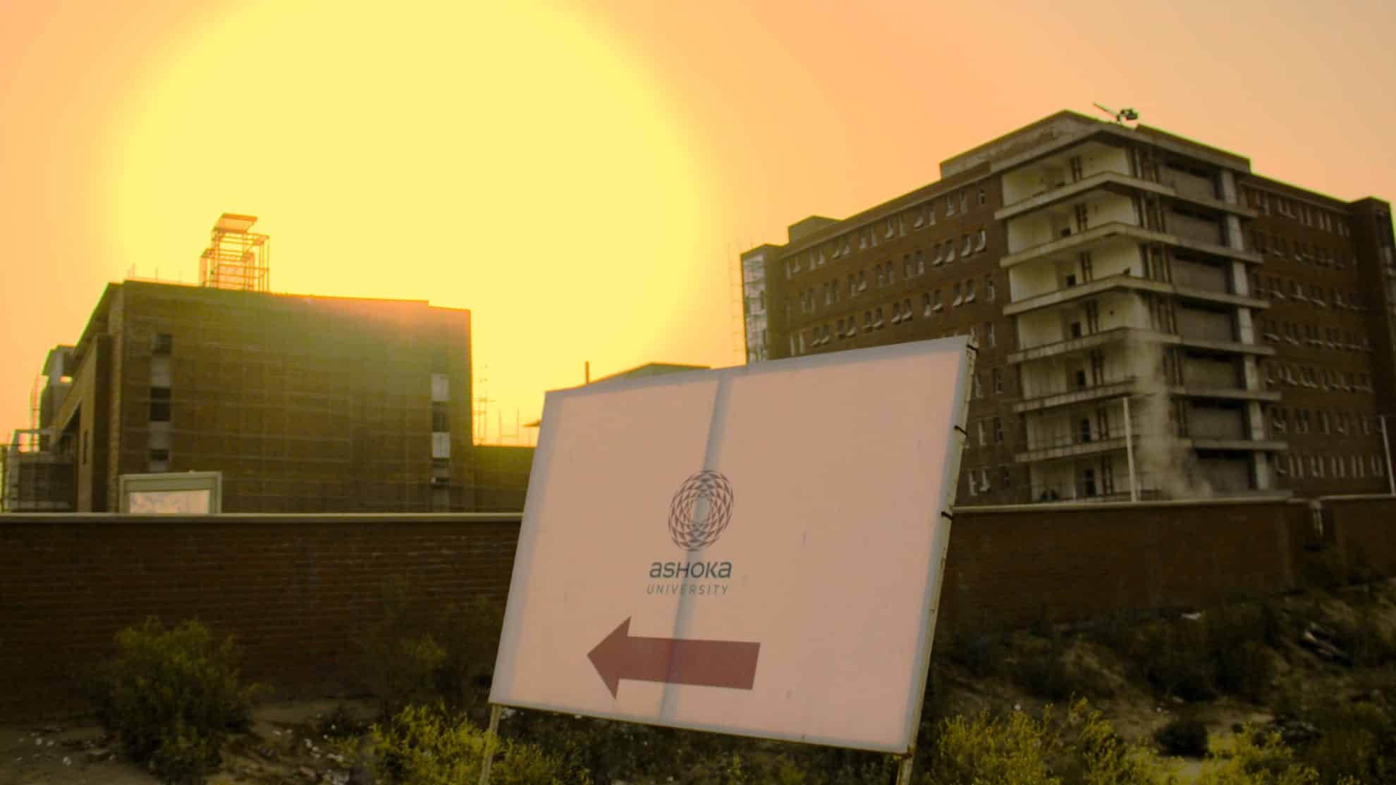 Sign with "Ashoka University" and arrow, in front of buildings at sunset, overgrown plants below.