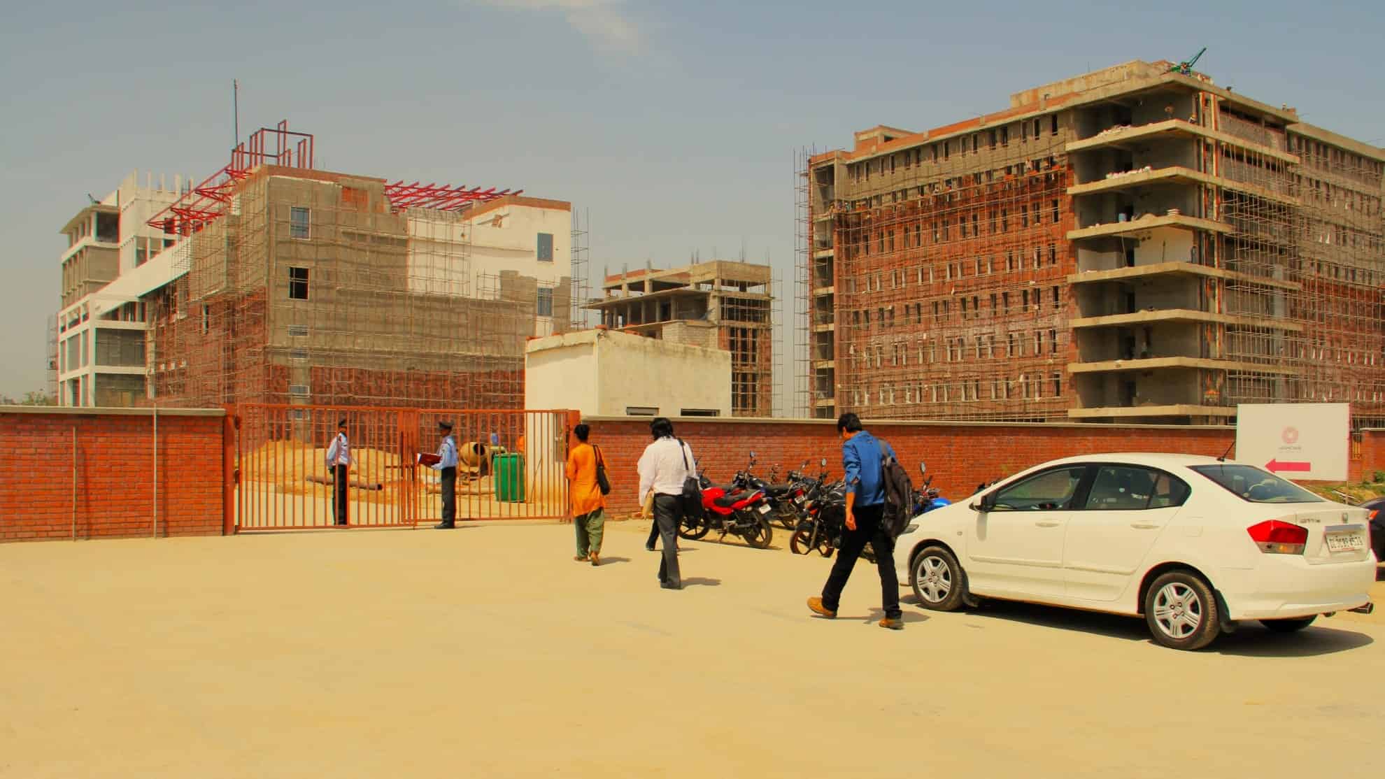 People walking near parked vehicles, red brick wall, and under-construction buildings in the background.