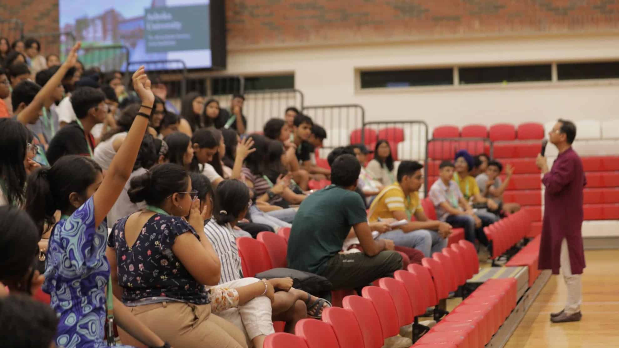 Audience seated in red chairs, some raising hands, speaker in purple attire addressing them.