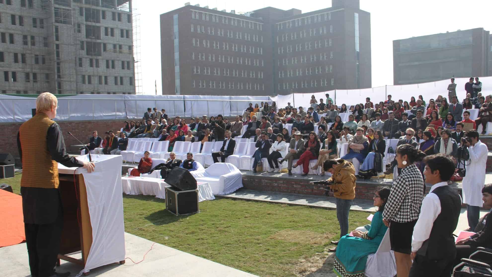 Speaker at a podium addressing an outdoor audience seated on rows of white chairs, with buildings behind.
