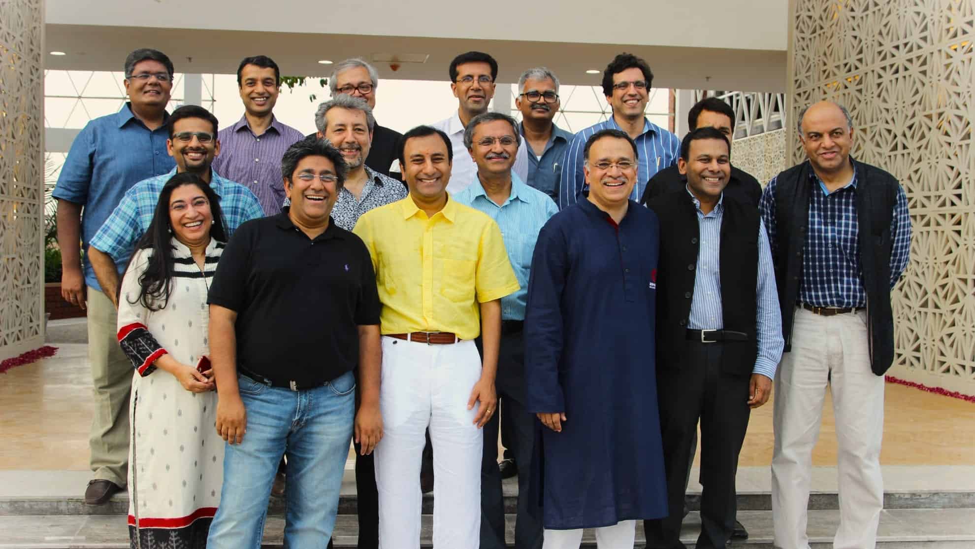 Group of 16 people smiling, dressed in casual and traditional attire, standing outdoors.