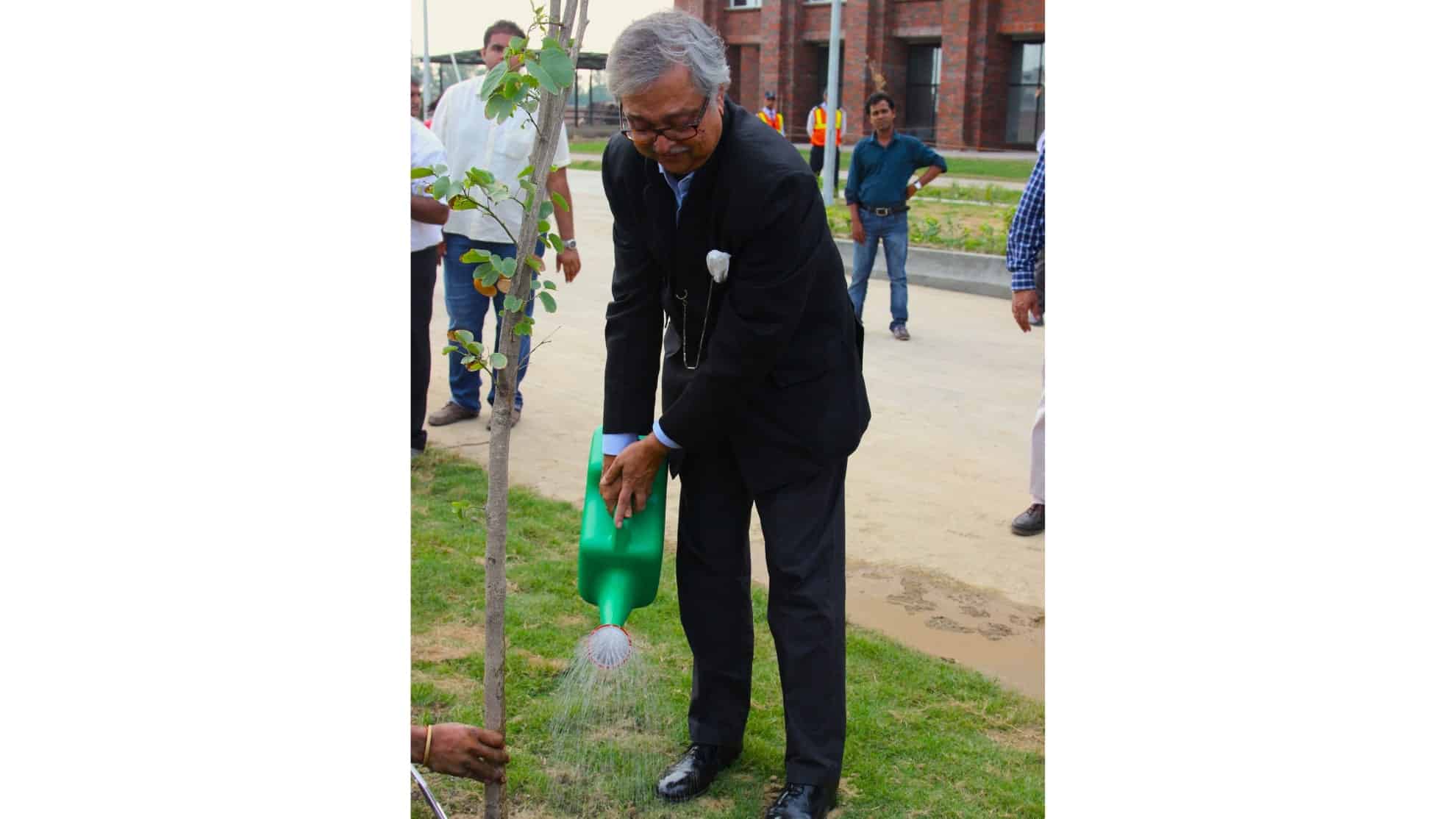 Man in a suit watering a small tree with a green watering can, surrounded by onlookers outdoors.