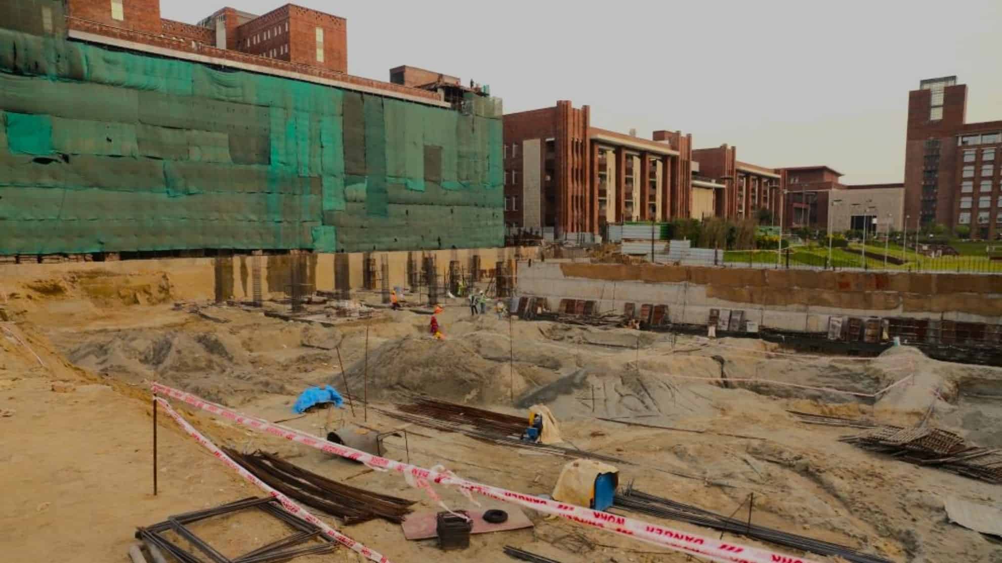 Construction site with excavation work, safety tape, workers, and adjacent red-brick buildings.