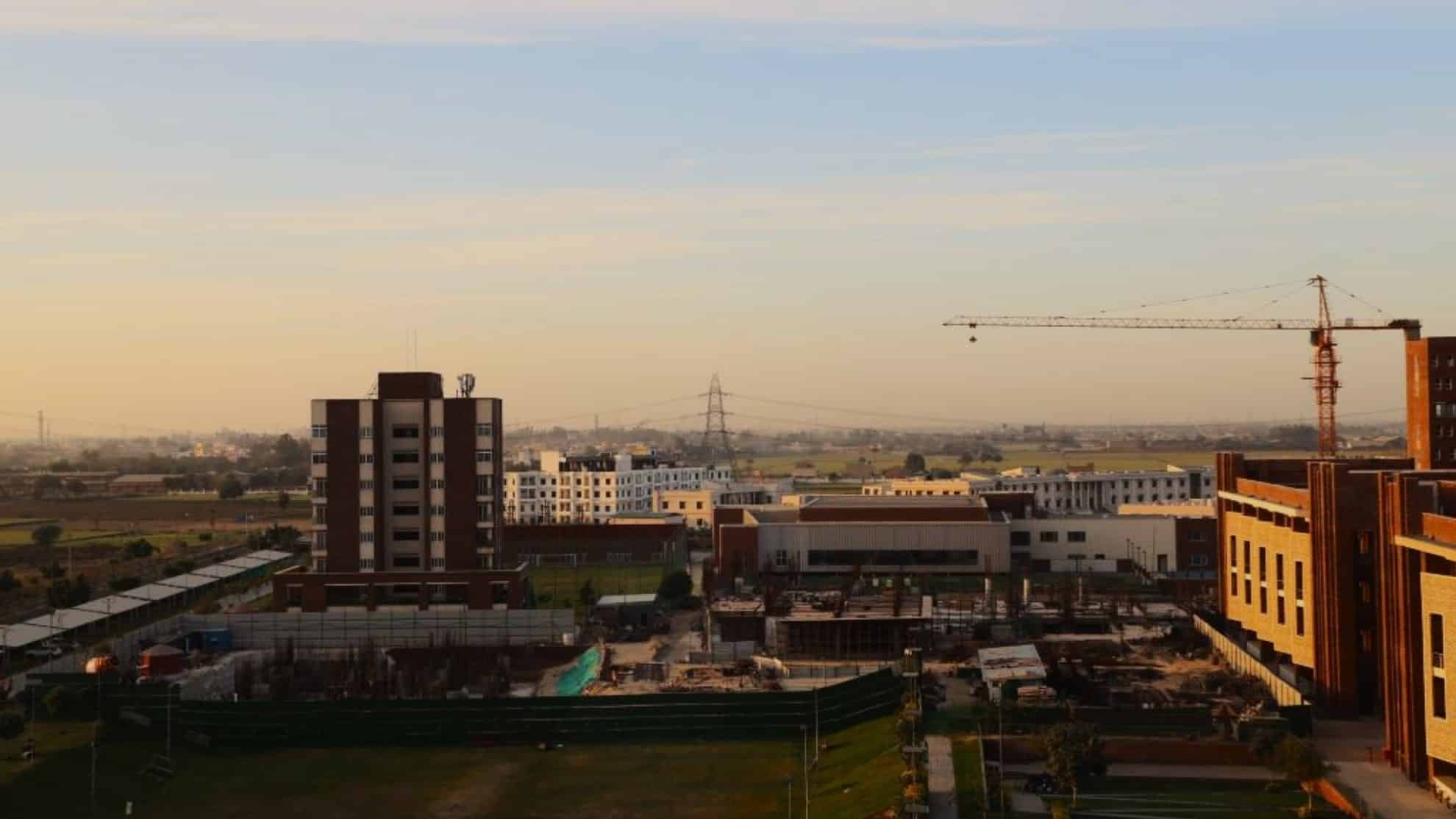 Buildings, construction site, crane, open fields, and power lines under a clear sky at sunset.