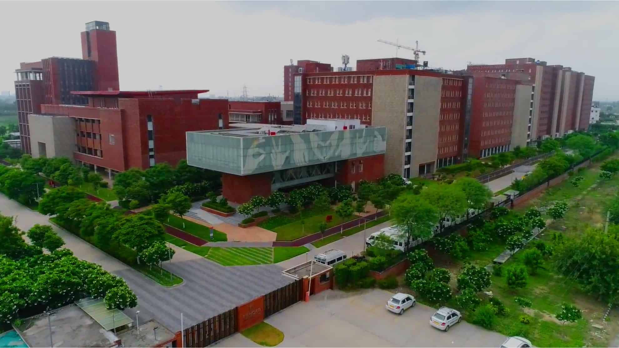 Red-brick campus buildings surrounded by green trees, roads, and parked cars under a cloudy sky.