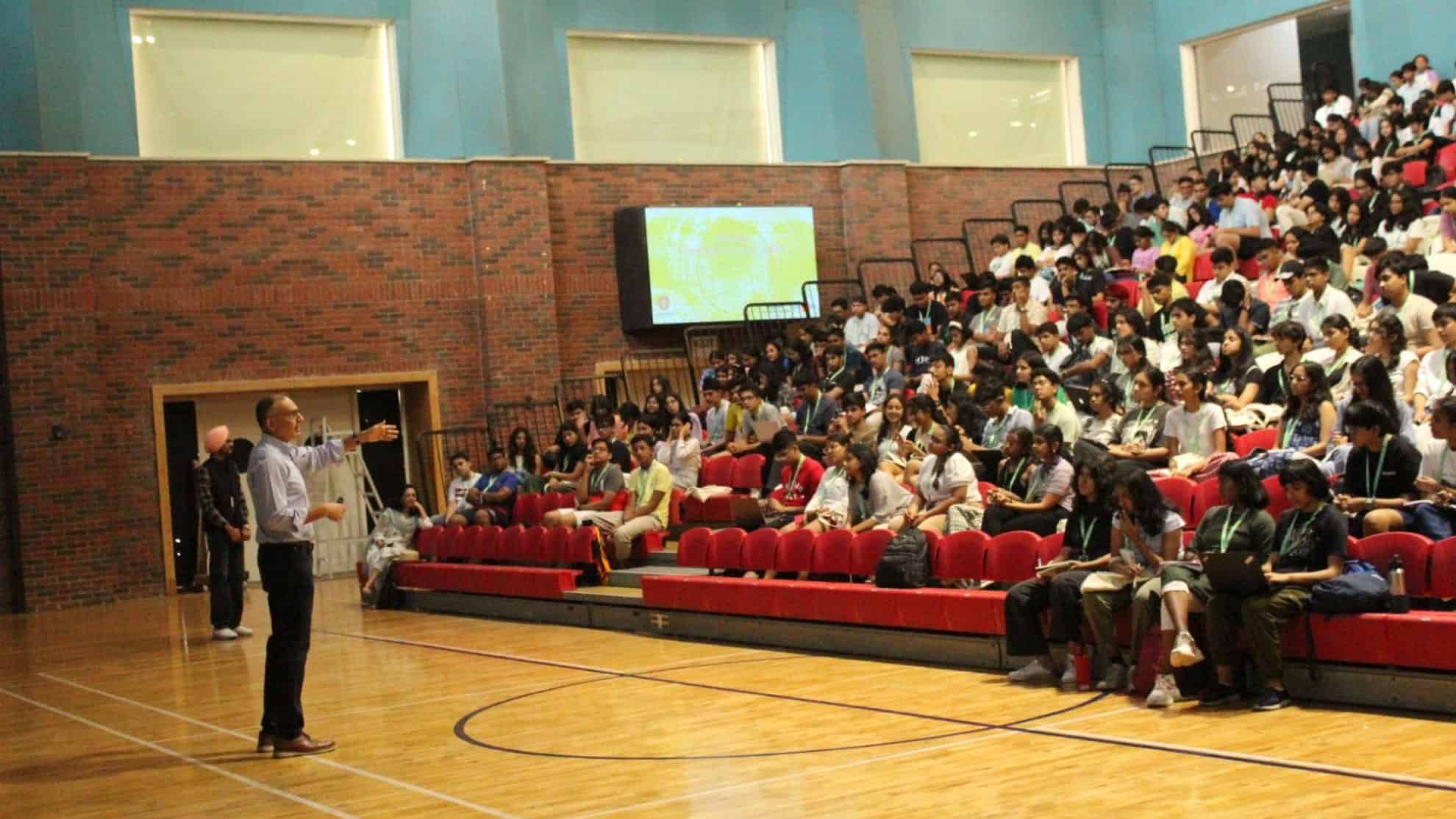 Speaker addressing a seated audience in a gymnasium with brick walls and a large screen behind.