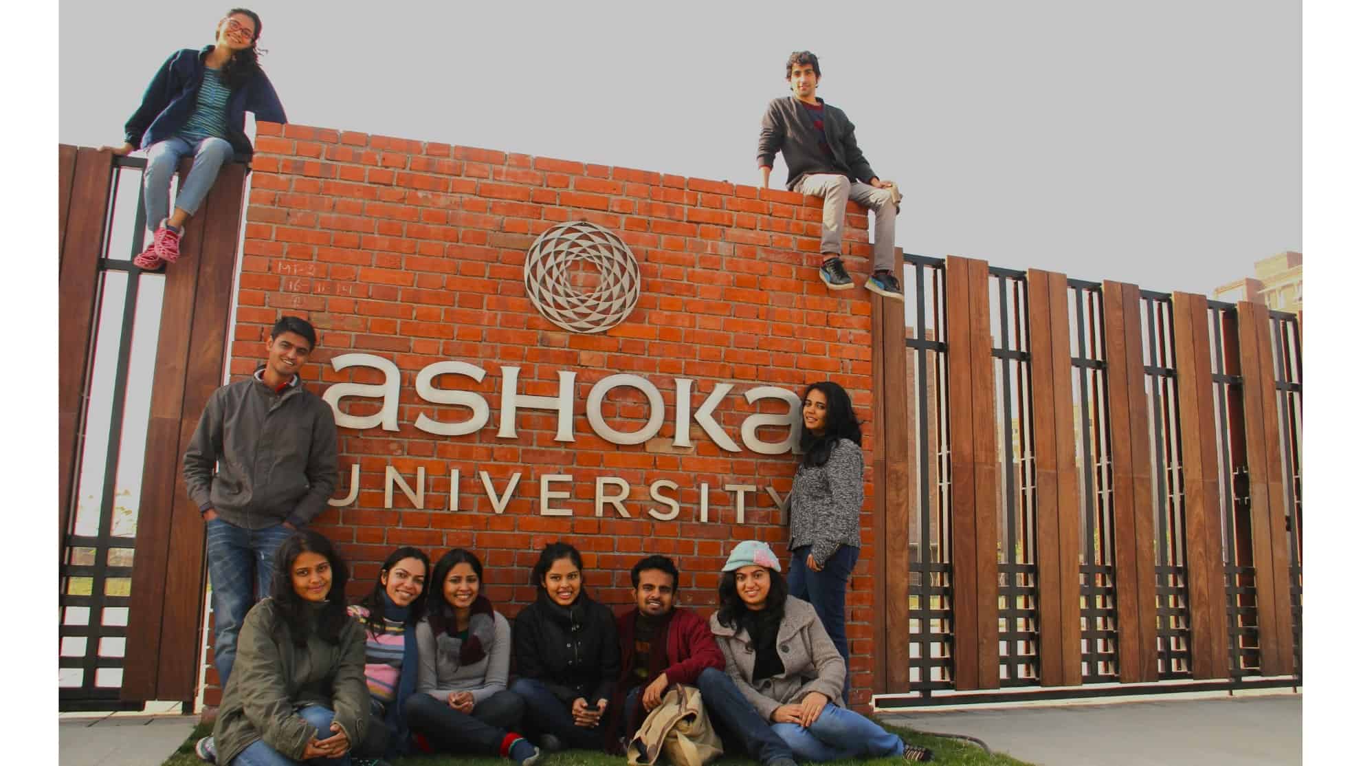 Group of students posing near Ashoka University brick wall and gate, with two sitting atop the wall.